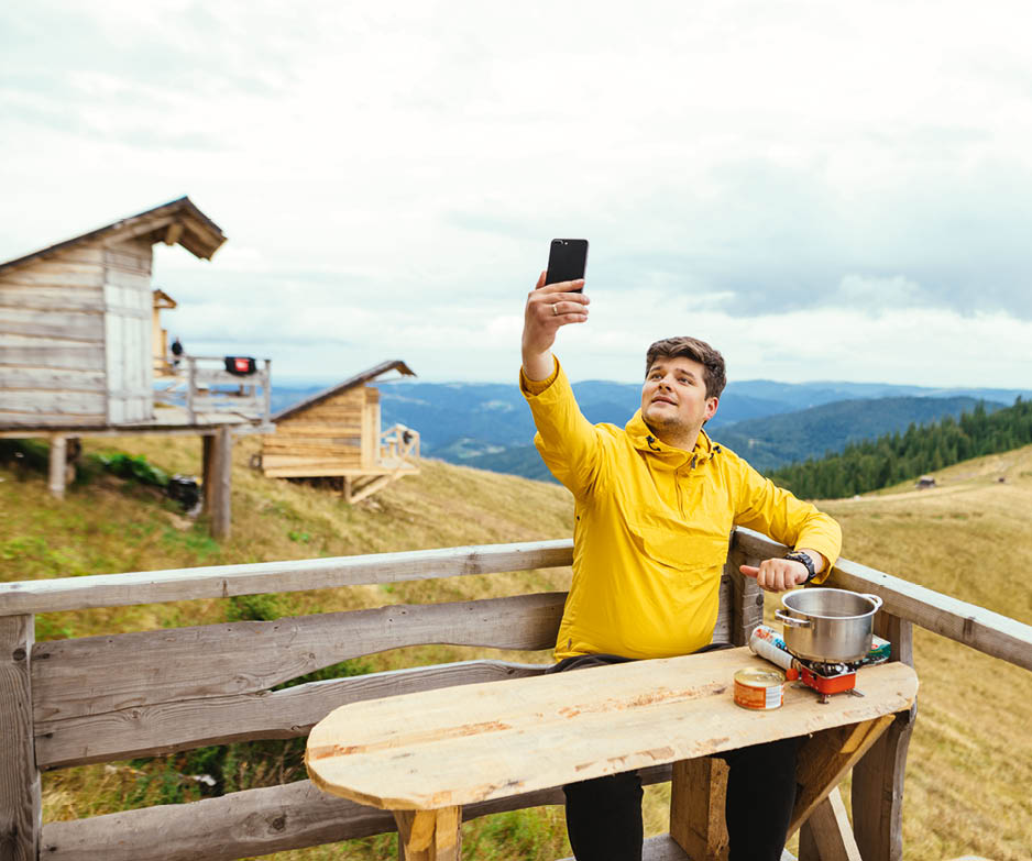 Positive man is sitting on a bungalow terrace at a table in the mountains, preparing food and using a smartphone with a cheerful face.
