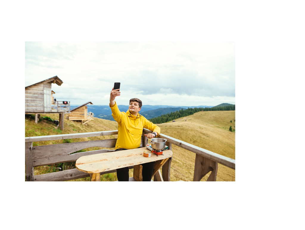 Positive man is sitting on a bungalow terrace at a table in the mountains, preparing food and using a smartphone with a cheerful face.
