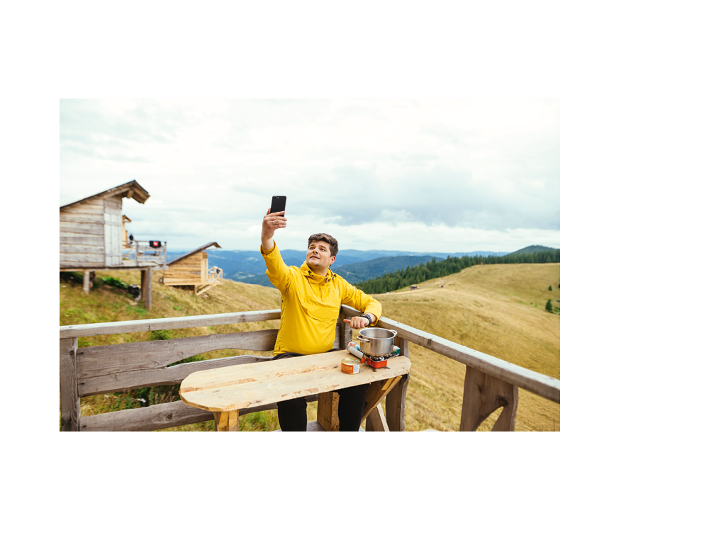 Positive man is sitting on a bungalow terrace at a table in the mountains, preparing food and using a smartphone with a cheerful face.